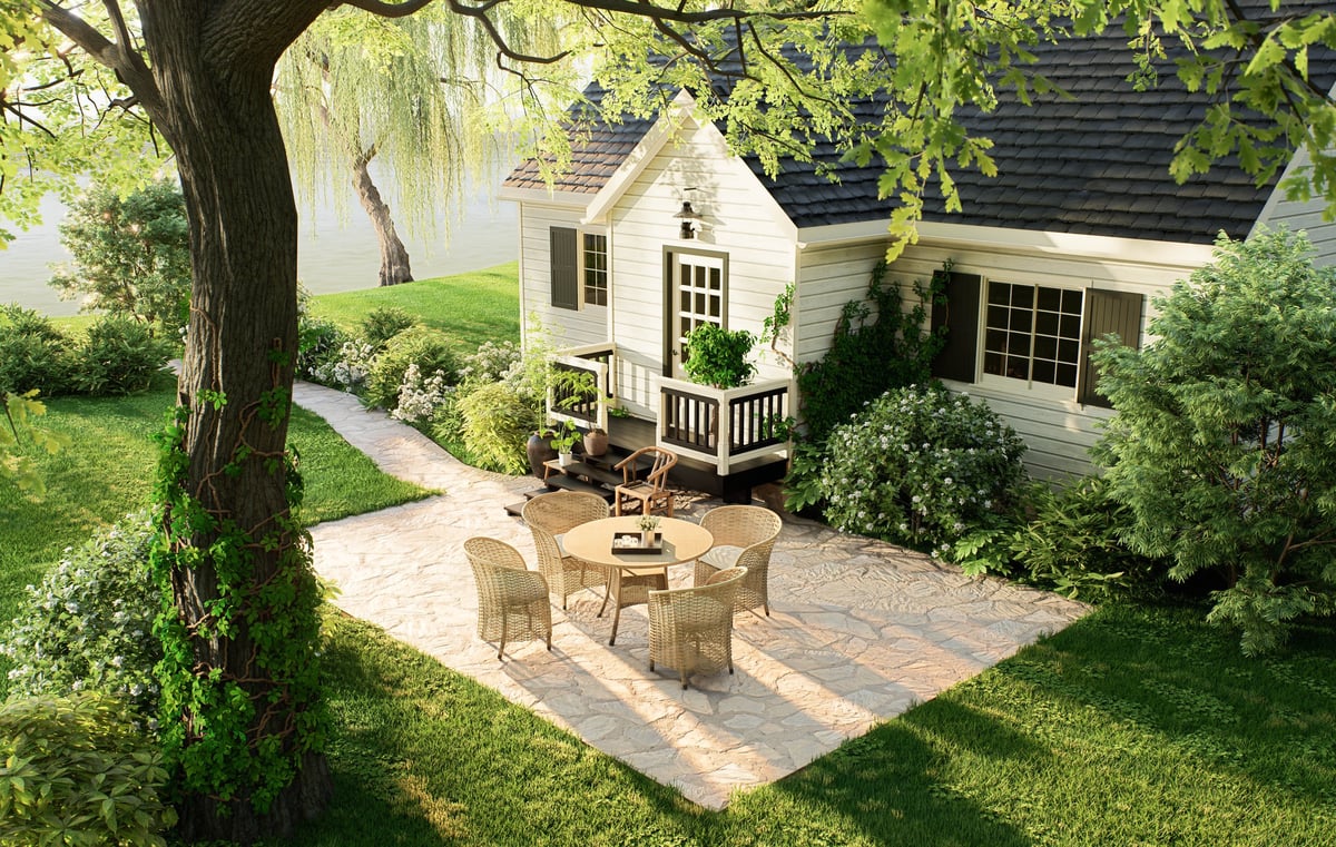 Charming country house backyard featuring a stone paved patio with a round wicker table and chairs, surrounded by a green lawn, lush trees, and a lake nearby.