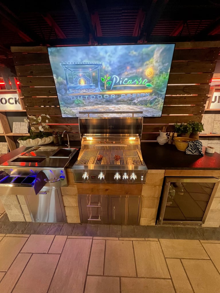Outdoor kitchen display with built-in grill, sink, and flat-screen TV mounted above showing Picasso Outdoor Design logo