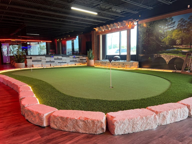 Indoor miniature golf course with green putting surface, pink stone border, red flooring, and screens displaying golf scenes